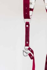 Detail shot of the lower section of the red leather camera harness showing the branded "GENUINESTRAP" metal connector, silver rivets, a snap shackle clip, and a leather safety tether strap.