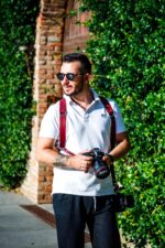 Front view of a photographer wearing the red leather dual camera harness over a white polo shirt, holding a Canon DSLR camera with a second camera hanging at his side, photographed outdoors against greenery and a brick wall.