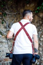 Rear view of a photographer wearing the red leather camera harness, showing the X-shaped cross-back design with silver buckle adjustments, two DSLR cameras hanging at each hip.