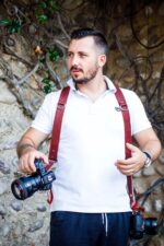 Front view of a photographer wearing the red leather dual camera harness, holding two Canon DSLR cameras at his sides, showing the shoulder straps with D-rings and the comfortable over-the-shoulder fit.