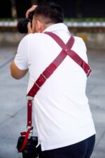 Rear action shot of a photographer using the red leather camera harness while shooting, with one camera raised to eye level and the second camera secured at his lower back, demonstrating the X-back strap design in use.