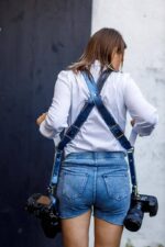Woman wearing Royal Blue Metallic leather camera harness viewed from behind, white long-sleeve shirt and blue denim shorts, dual professional cameras attached to metal carabiner clips