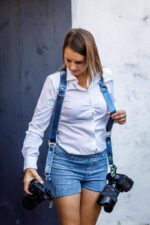 Person wearing Royal Blue Metallic leather camera harness in white shirt and blue jeans, holding camera with metal carabiner clip visible