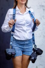 Woman wearing Royal Blue Metallic leather camera harness, white long-sleeve shirt and blue denim shorts, carrying dual DSLR cameras