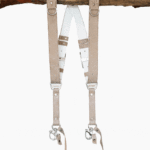 Sand-colored vintage leather dual camera harness with silver hardware, hanging from a rustic wooden branch against a white background, showing the full front view with two shoulder straps, a white leather back panel, branded "GENUINESTRAP" metal connectors, and snap hook attachments at each end.