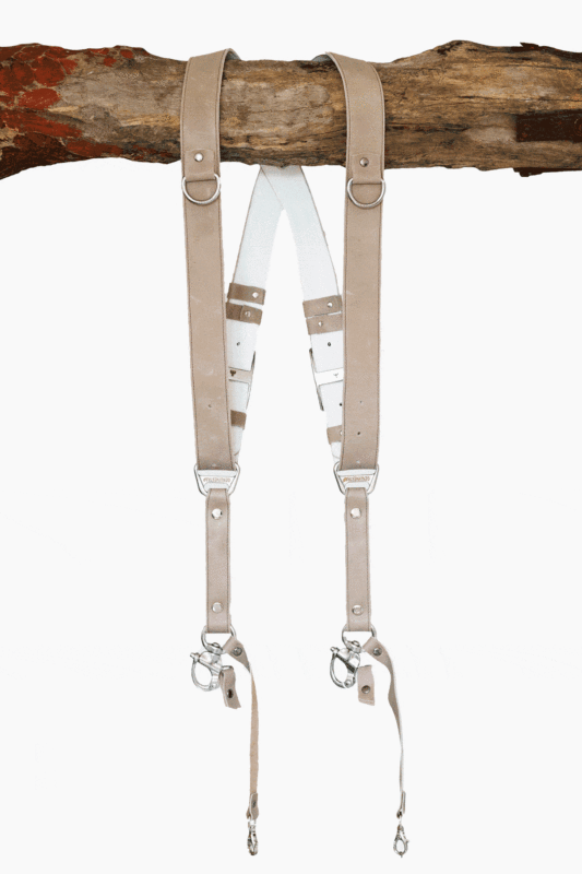 Sand-colored vintage leather dual camera harness with silver hardware, hanging from a rustic wooden branch against a white background, showing the full front view with two shoulder straps, a white leather back panel, branded "GENUINESTRAP" metal connectors, and snap hook attachments at each end.