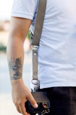 Close-up side view of a photographer wearing the sand-colored leather camera harness, showing the strap running from shoulder to hip with the branded "GENUINESTRAP" metal connector, D-ring, snap shackle clip, and a camera attached at waist level.