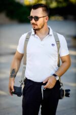 Front view of a photographer wearing sunglasses and the sand-colored leather dual camera harness over a white polo shirt, with two cameras hanging at his hips, showing the shoulder straps with D-rings and the overall relaxed fit of the harness.