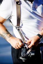 Close-up of a photographer's hands attaching a camera to the sand-colored leather harness using the snap shackle clip, showing the branded metal connector, riveted leather strap, and the quick-release mechanism in action.