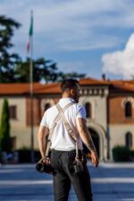 Rear view of a photographer wearing the sand-colored leather dual camera harness over a white polo shirt, walking through an Italian piazza with two cameras hanging at his hips, showing the X-shaped cross-back strap design.