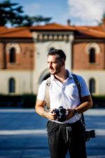 Front view of a photographer wearing the sand-colored leather camera harness over a white polo shirt, holding a camera at waist level with a second camera hanging at his side, photographed outdoors in front of a historic Italian building.