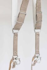 Close-up detail of the lower section of the sand-colored leather camera harness showing both strap ends with silver adjustable buckles, branded "GENUINESTRAP" metal connectors, riveted construction, snap shackle clips, and leather safety tethers.