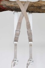 Back view of the sand-colored leather camera harness hanging from a rustic wooden branch, displaying the X-cross strap configuration, silver adjustable buckles, branded metal connectors, and dual snap shackle attachment points at the bottom.