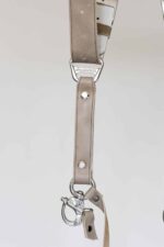 Detail shot of a single strap end of the sand-colored leather camera harness showing the branded "GENUINESTRAP" metal connector, silver rivet, D-ring, snap shackle clip, and leather safety tether against a light background.