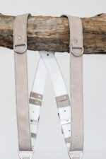 Close-up of the upper portion of the sand-colored leather camera harness hanging from a wooden branch, highlighting the silver D-rings, white leather backing with riveted leather keepers, and the branded "GENUINESTRAP" metal connectors at the junction points.