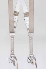 Front mid-section detail of the sand-colored leather camera harness showing both straps with the white leather back panel, branded "GENUINESTRAP" connectors, silver rivets, and snap shackle clips with leather safety tethers at the bottom.