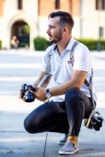 Photographer crouching outdoors while wearing the sand-colored leather dual camera harness, holding one camera in his hands with a second hanging at his side, demonstrating the harness's flexibility and comfort during active shooting.