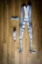 Flat lay on a wooden floor showing the complete silver glitter leather camera harness set, including the dual camera harness with white leather backing and snap shackle clips, a matching glitter leather bow tie, and a matching glitter leather belt with silver buckle.