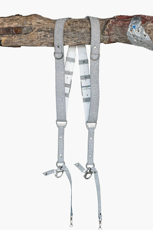 Silver glitter leather dual camera harness hanging from a rustic wooden branch against a light background, showing the full front view with two sparkle-finish shoulder straps, white leather back panel, D-rings, metal connectors, snap shackle clips, and lobster claw clasps at each end.