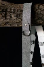 Close-up detail of the upper portion of the silver glitter leather camera harness hanging from a wooden branch against a dark background, highlighting the sparkle texture of the leather, the silver rivet, and the D-ring attachment point with the white leather backing visible behind.