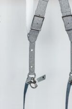Detail shot of a single strap junction on the silver glitter leather camera harness, showing the adjustable rectangular buckle, white leather backing, D-ring connector, riveted glitter leather strap, snap shackle clip, and leather safety tether against a light background.