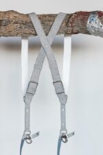 Back view of the silver glitter leather dual camera harness hanging from a rustic wooden branch, displaying the X-cross strap configuration, white leather backing, adjustable rectangular buckles, and dual snap shackle attachment points at the bottom.