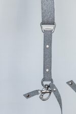 Close-up detail of the lower section of the silver glitter leather camera harness showing the branded "GENUINESTRAP" metal connector, riveted sparkle-finish strap, D-ring, snap shackle clip, and leather safety tether against a light grey background.