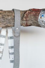 Close-up of the upper shoulder area of the silver glitter leather camera harness draped over a rustic wooden branch, highlighting the sparkle leather texture, silver rivet, D-ring, and the white leather back panel with glitter leather keepers visible alongside.