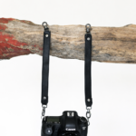 Black leather camera strap attached to camera and hanging from rustic wood branch on a light background.