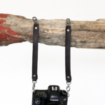 Black leather camera strap attached to camera and hanging from rustic wood branch on a light background.