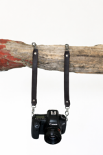 Black leather camera strap attached to camera and hanging from rustic wood branch on a light background.