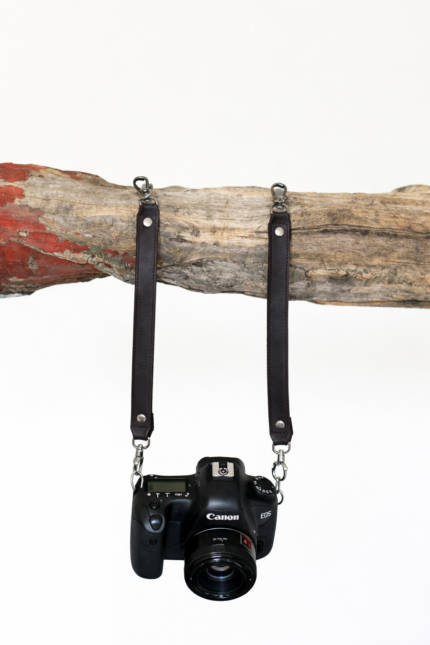 Black leather camera strap attached to camera and hanging from rustic wood branch on a light background.