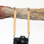 Light brown leather camera strap attached to camera and hanging from rustic wood branch on a light background.