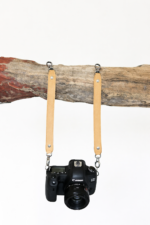 Light brown leather camera strap attached to camera and hanging from rustic wood branch on a light background.