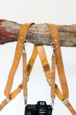 Pair of tan leather third camera harness extenders with metal clasps hanging on a wooden branch