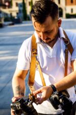 Photographer wearing tan leather camera harness extenders while adjusting gear outdoors