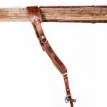 Full-length vintage brown leather camera strap with padded shoulder section, adjustable buckle holes, metal rings, and clasp hardware hanging from a rustic wooden beam on a light gray background.
