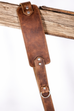 Vintage brown leather camera strap close-up with stitched shoulder pad, metal rivets, and ring detail hanging over a rustic wooden beam on a light gray background.