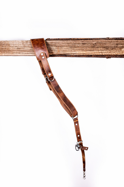 Full-length vintage brown leather camera strap with padded shoulder section, adjustable buckle holes, metal rings, and clasp hardware hanging from a rustic wooden beam on a light gray background.