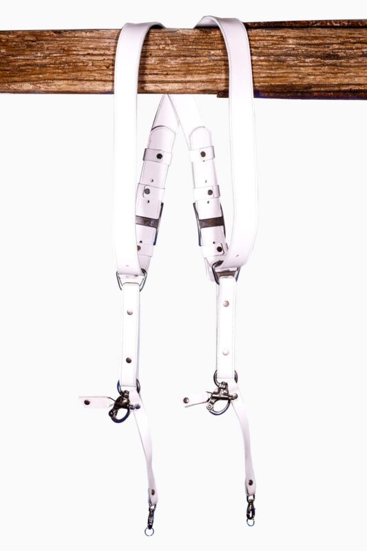 White leather dual camera harness hanging from a rustic wooden beam with adjustable straps and silver metal clips.