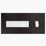 Top-down close-up of two leather desk mats side by side — one glossy medium brown and one matte dark brown — with a white wireless mouse resting on the darker mat, on a light stone surface.