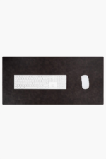 Top-down close-up of two leather desk mats side by side — one glossy medium brown and one matte dark brown — with a white wireless mouse resting on the darker mat, on a light stone surface.