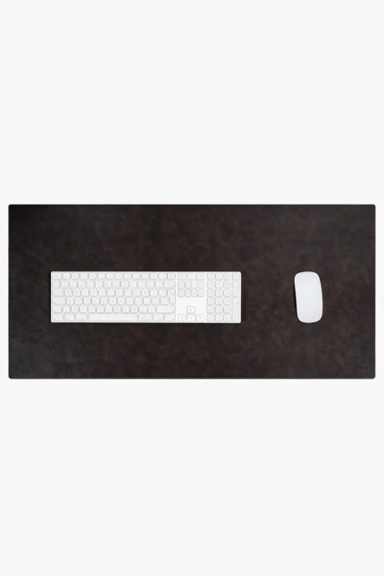 Top-down close-up of two leather desk mats side by side — one glossy medium brown and one matte dark brown — with a white wireless mouse resting on the darker mat, on a light stone surface.
