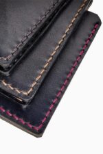 Close-up detail of the corner of multiple black leather bifold wallets stacked together, showing hand-stitching in three different thread colors: dark brown, natural tan, and fuchsia pink, against a white background.