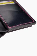 Close-up detail of the interior lower corner of a black leather bifold wallet, highlighting the card pocket and the precise fuchsia pink hand-stitching along the edges, against a white background.