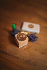 Custom wood stamp with engraved circular laurel wreath design on a wooden tabletop, with ink pad and dried lavender softly blurred in the background.