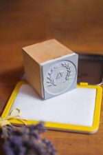 Custom wood stamp with engraved laurel wreath design beside an ink pad on a wooden tabletop with dried lavender.