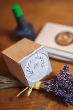 Close-up of a custom wood stamp with engraved logo resting on a bundle of dried lavender, with ink pad in the background on a wooden tabletop.