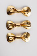 Three gold metallic leather bow ties neatly stacked vertically on a white background, showing consistent craftsmanship and the luminous herringbone texture across multiple pieces.
