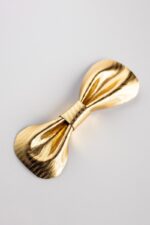 Gold metallic leather bow tie photographed at an angle on a light grey background, highlighting the herringbone embossed texture and gathered center knot.